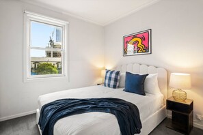 Une chambre d’hôtel avec un lit, une fenêtre donnant sur des immeubles, une œuvre d’art accrochée au mur et une lampe de chevet.