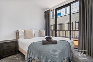 A modern hotel room with a large bed, a nightstand, and a view of a balcony with a metal fence. - Riverside Family Getaway (Melbourne)