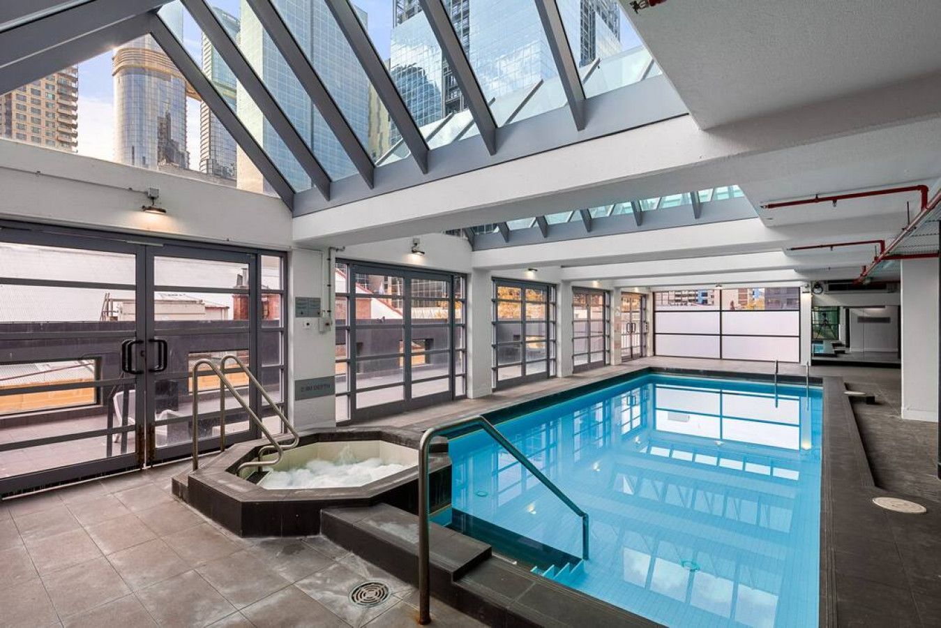 A modern indoor swimming pool with a glass ceiling, a hot tub, and a view of the cityscape.