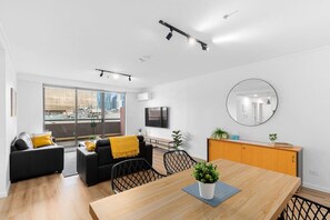 A modern living room with a sofa, a dining table with a plant, and a large window offering a city view. - 2 Bedroom in the Paris End (Melbourne)