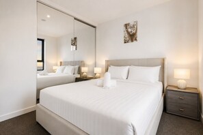 A hotel room with a large bed, a bedside table with a lamp, a mirror, and a painting on the wall. - Vibrant Southbank 2 Bedroom (Southbank)