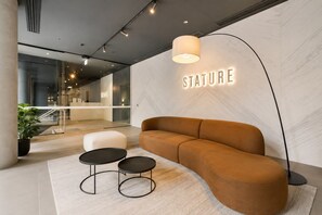Moderni aula, jossa on sohva, kaksi pyöreää pöytää, lattiavalaisin ja kyltti, jossa lukee »STATURE».