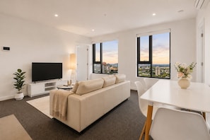A modern living room with a sofa, TV, and dining area, featuring large windows with a city view. - Vibrant Southbank 2 Bedroom (Southbank)