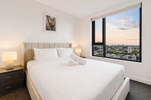 A hotel room with a large bed, a nightstand, a lamp, a framed picture on the wall, and a window with a city view. - Vibrant Southbank 2 Bedroom (Southbank)