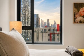 A room with a large window overlooking a cityscape, a lamp, a painting, and a rolled towel on the bed. - Luxury Southbank Skyline Stay (Southbank)