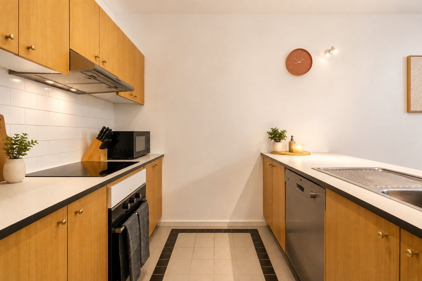 A compact kitchen with wooden cabinets, a microwave, a sink, and a clock on the wall.