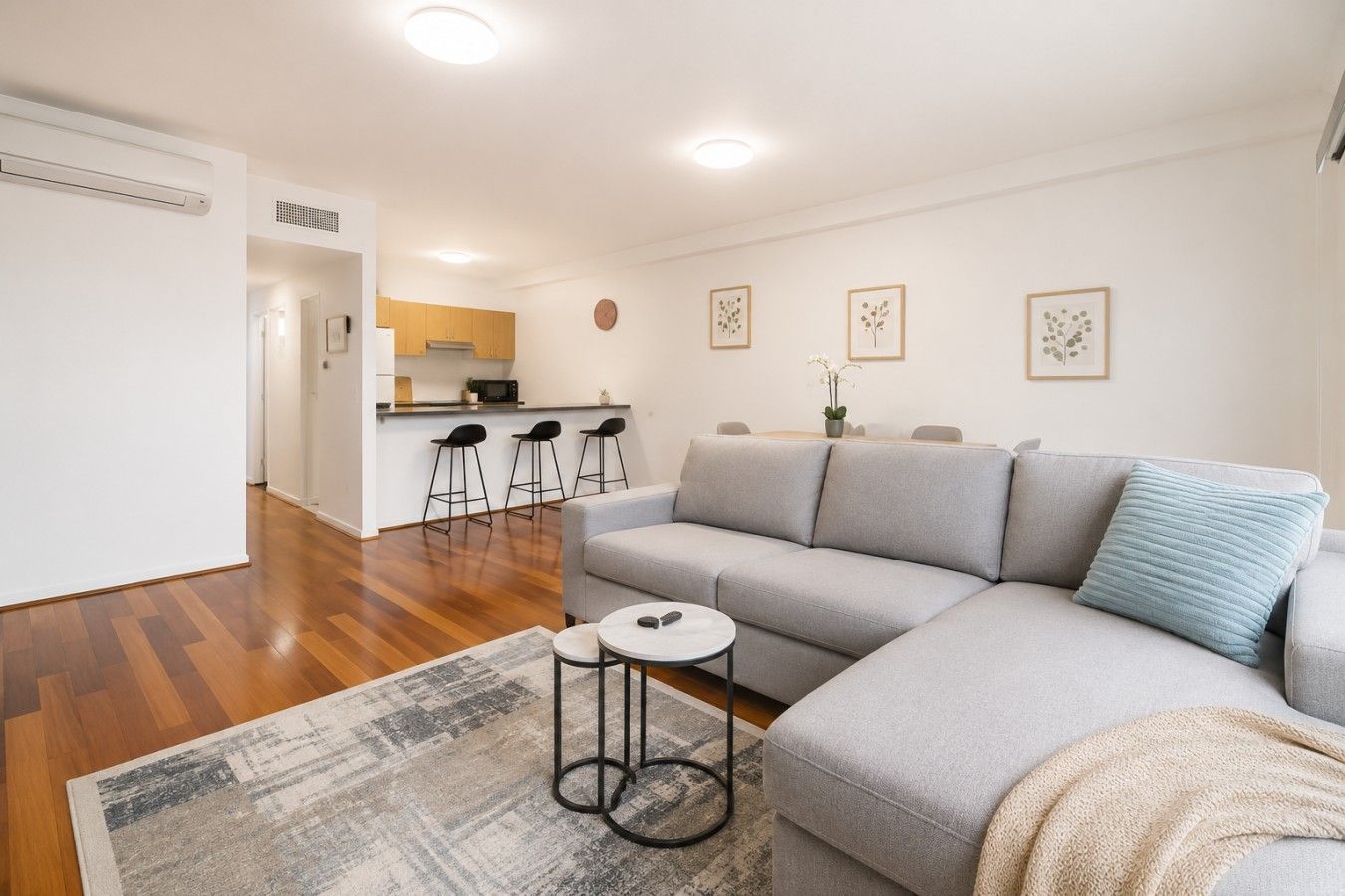 A modern living room with a grey sofa, a small round table, and a coffee table. There is a kitchen area in the background with a microwave and bar stools.