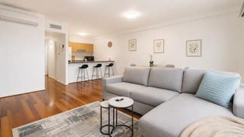 A modern living room with a grey sofa, a small round table, and a coffee table. There is a kitchen area in the background with a microwave and bar stools.