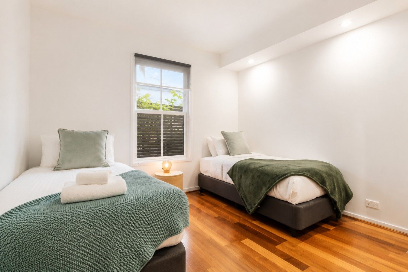 A bedroom with two beds, a window with blinds, a bedside table with a lamp, and a wooden floor.