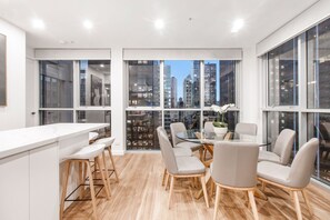A modern kitchen with a white countertop, wooden stools, and a glass dining table with grey chairs. - City 4 Bedroom Penthouse (Melbourne)