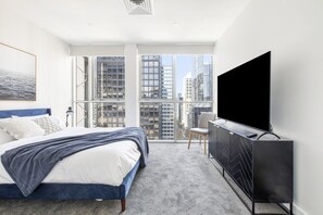 A modern bedroom with a large bed, a TV, and a view of city buildings. - City 4 Bedroom Penthouse (Melbourne)