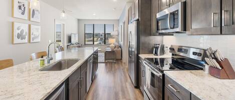 A modern kitchen with stainless steel appliances, a marble countertop, and a view to the living area.