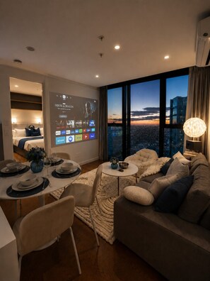 Sala moderna com vista para a cidade, área de jantar com mesa posta para dois e uma grande tela exibindo um filme.