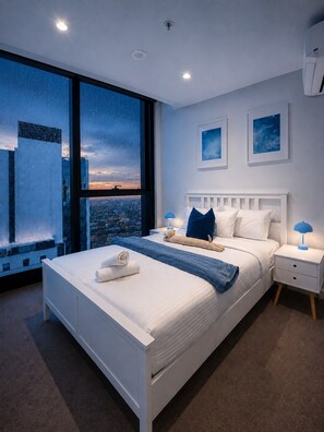 Une chambre moderne avec un grand lit, une table de chevet, une fenêtre donnant sur la ville et deux tableaux encadrés accrochés au mur.