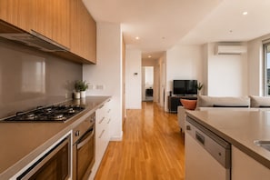 A modern kitchen with a stove, oven, and dishwasher, leading to a living area with a sofa and a TV. - Bay View 2 Bedroom Apartment (Port Melbourne)