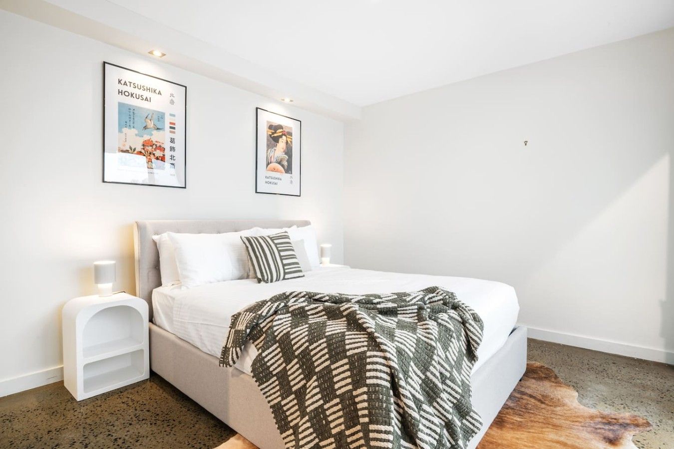A bedroom with a bed, a nightstand, two framed posters, and a rug.