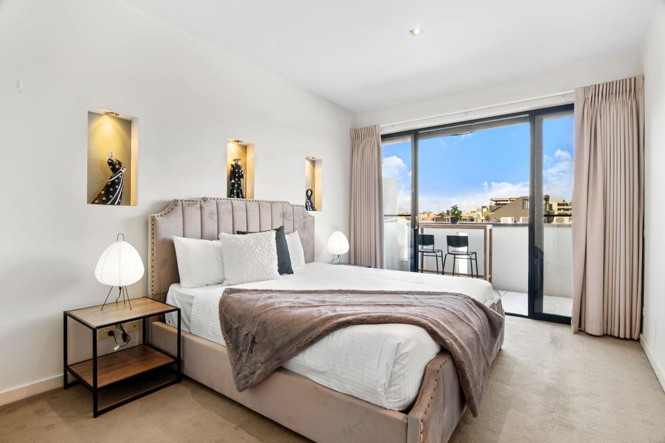 A modern bedroom with a large bed, bedside lamps, and a view of the city through a balcony.
