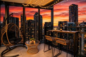A rooftop terrace with a city skyline view, a hanging chair, two bar stools, and a table with a bottle and glasses. - Skyline Sunset 2 Bedroom (Southbank)