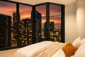 A bedroom with a cityscape view through floor-to-ceiling windows, a bed with white linens, and a textured throw blanket. - Skyline Sunset 2 Bedroom (Southbank)