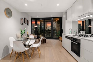 A modern kitchen with a dining area, a built-in stove, and a view of the cityscape through large windows. - Skyline Sunset 2 Bedroom (Southbank)