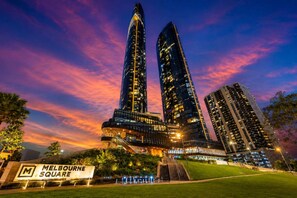 A high-rise building complex with a prominent 'Melbourne Square' sign, surrounded by greenery and under a twilight sky. - Skyline Sunset 2 Bedroom (Southbank)