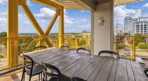 A balcony with a wooden table and chairs, a yellow railing, and a city view. - Landing | Amazing Studio, Clubhouse, Gym (Charlotte)