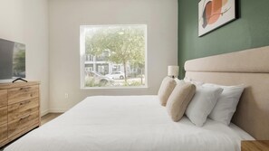 A bedroom with a large window, a bed with white bedding, a wooden dresser, and a wall-mounted lamp. - Landing | Spectacular 1BD in North Kissimmee (Kissimmee)