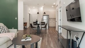 Una sala moderna con sofá, una mesa de centro con adornos, un comedor con sillas y una mesa, una cocina con armarios y un refrigerador, y un televisor montado en la pared.