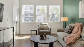 A living room with a sofa, armchair, coffee table, and a TV on the left. There are three windows providing natural light. - Landing | Beautiful 2BD in North Kissimmee (Kissimmee)
