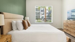 A bedroom with a bed, pillows, a nightstand, a dresser, a TV, and a window with a view of buildings and a palm tree. - Landing | Modern 2BD in North Kissimmee (Kissimmee)