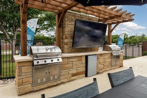 An outdoor barbecue area with a TV, two grills, and a stone-built structure. - Landing | Beautiful 2BD, Gym, Courtyard (Allen)