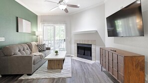 A living room with a sofa, coffee table, fireplace, TV, and a ceiling fan. - Landing | Chic 2BD, Courtyard, Gym (Allen)