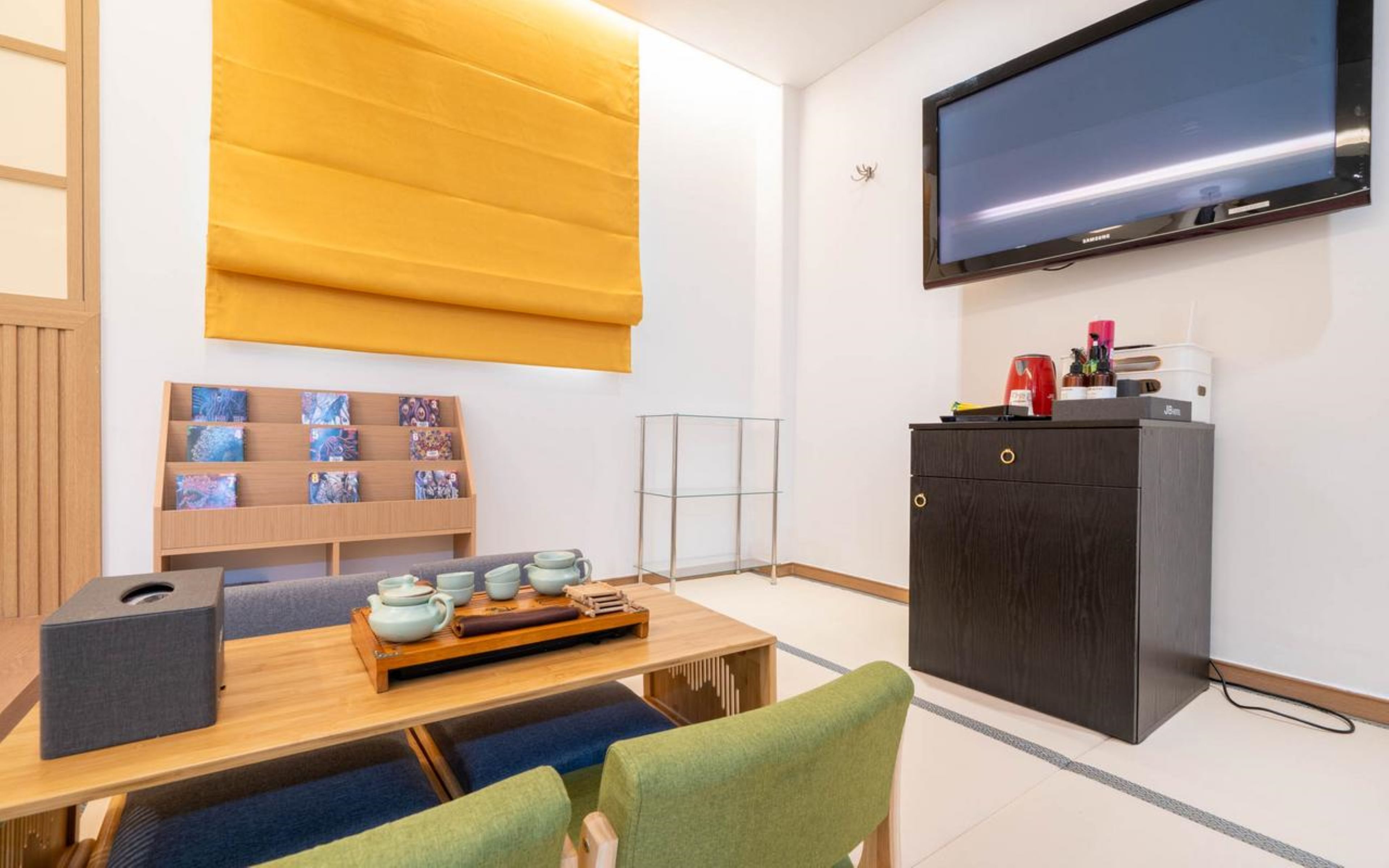 A room with a television mounted on the wall, a wooden table with a tea set, a bookshelf with books, and a cabinet with a coffee machine.