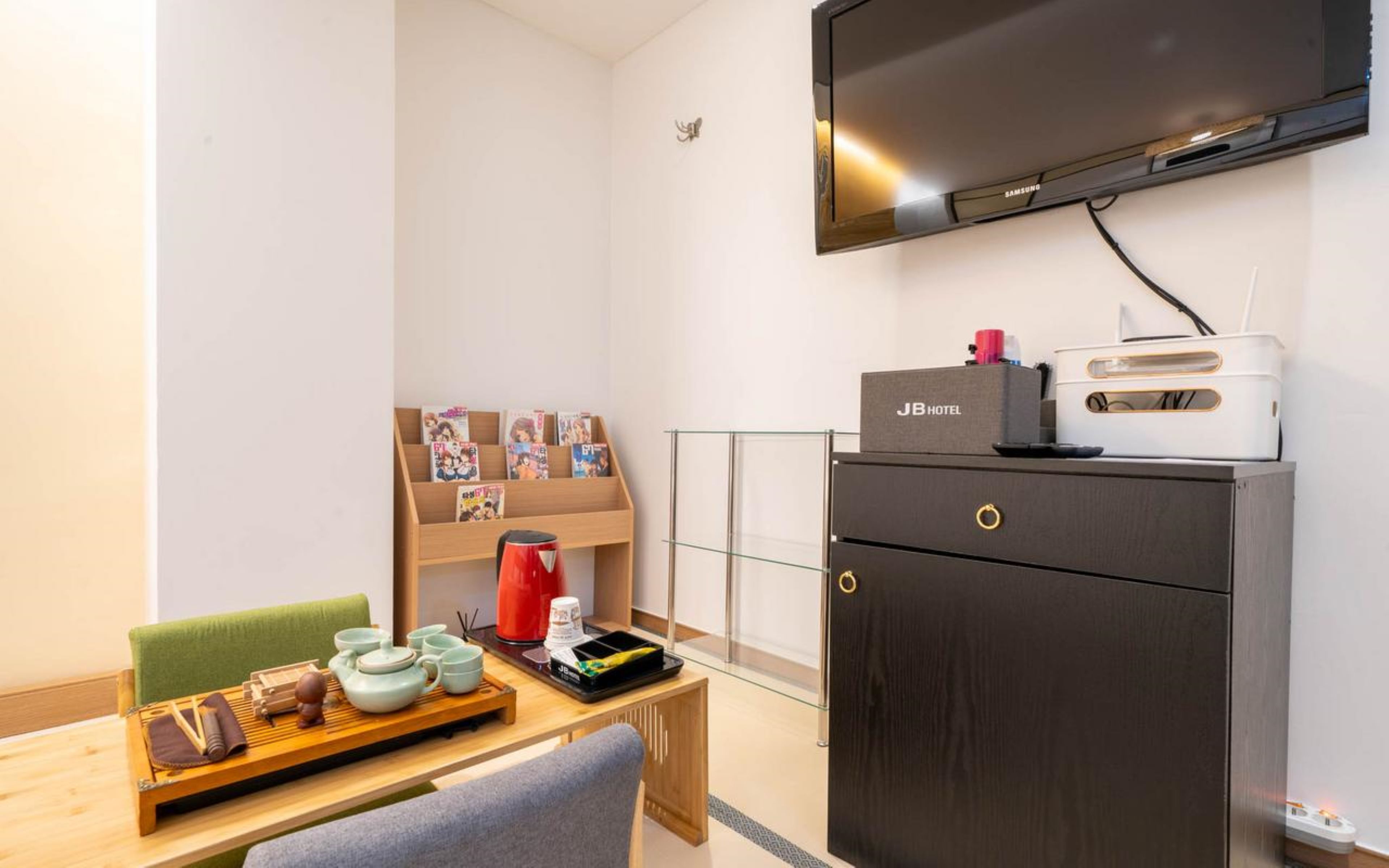 A hotel room with a flat-screen TV mounted on the wall, a wooden table with a tray of tea utensils, a red kettle, and a bookshelf with magazines.