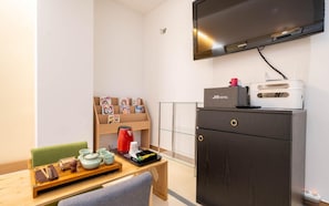Habitació d'hotel amb televisió de pantalla plana penjada a la paret, una taula de fusta amb una bandeja de vaixelles per a te, una paperera vermella i una prestatgeria amb revistes.