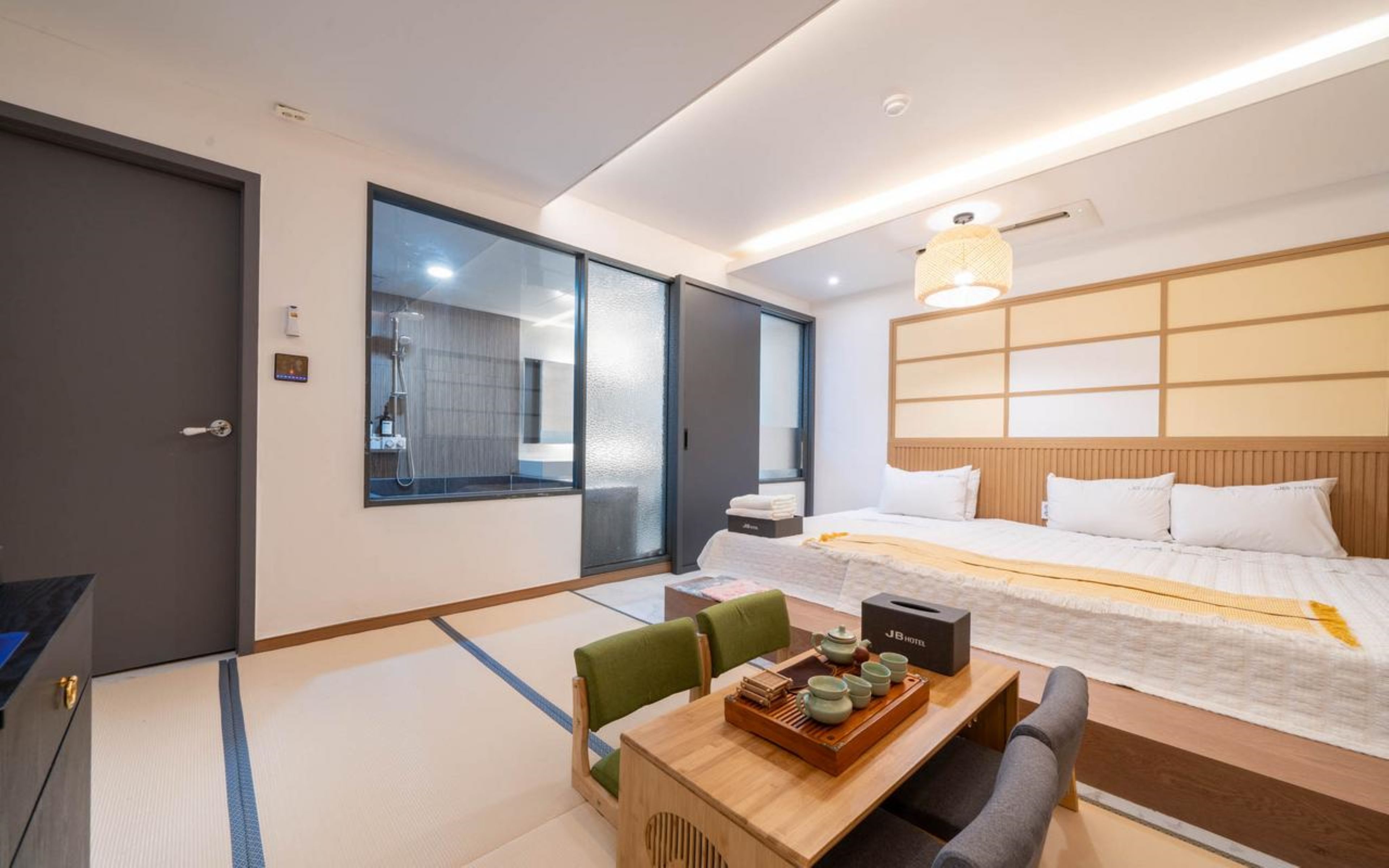 A hotel room with a bed, a desk with a tray of tea, a chair, a sliding door, and a large window.