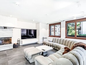 A modern living room with a fireplace, a sectional sofa, and a large window offering a view of buildings and greenery. - Anges Zermatt by Interhome (Zermatt)