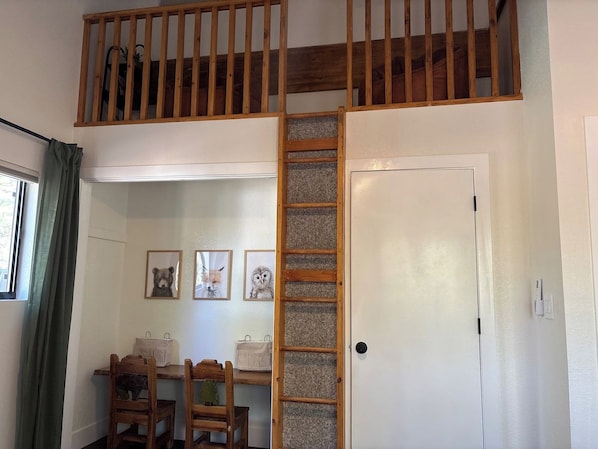 A room with a loft bed, a wooden ladder, a desk with chairs, and framed pictures on the wall.