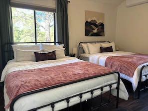 A room with two beds, each with a white mattress and a red blanket. There is a window with curtains, a wall-mounted artwork, and a bedside table with a lamp.