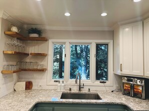 A kitchen with a granite countertop, a double sink, wooden shelves with glassware, and a window with a view of trees. - Lafayette Hills 3BR Private Family Getaway Near Walnut Creek (Lafayette)