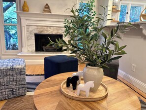 Une pièce avec une cheminée, un pouf bleu, une table ronde en bois agrémentée d’objets décoratifs, et une vue sur les arbres par les fenêtres.