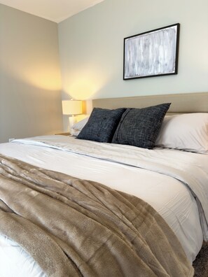 Un lit bien fait, recouvert d’une couverture beige et agrémenté de deux oreillers, une lampe de chevet et un tableau abstrait encadré accroché au mur.