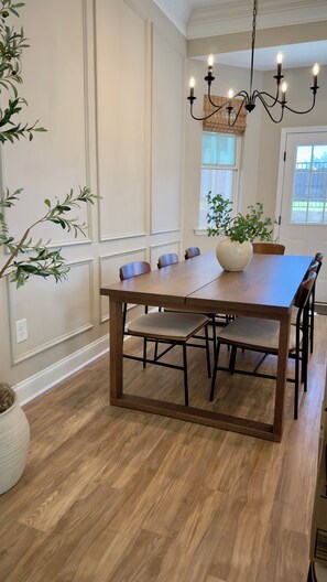 Een eethoek met een houten tafel, zes stoelen, een kroonluchter en potplanten.