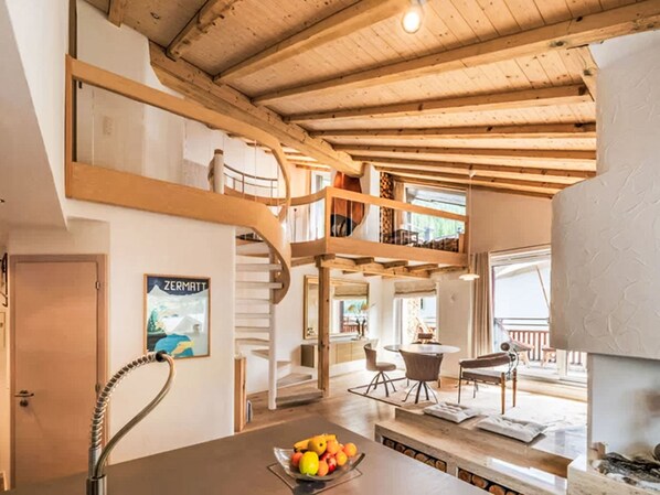 Ein geräumiger Innenraum mit hoher Decke, Holzbalken, einer Treppe, einem Essbereich und einer Obstschale auf der Arbeitsfläche.