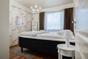 A bedroom with a bed, a bedside table, a chandelier, a window with curtains, and a patterned wallpaper. - Ounasrinne 3BR House, Sauna By Lapland Host (Rovaniemi)
