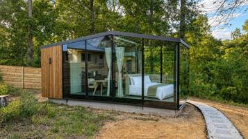 Une cabane moderne aux parois entièrement vitrées, avec un lit et un coin repas visibles à travers la vitre.
