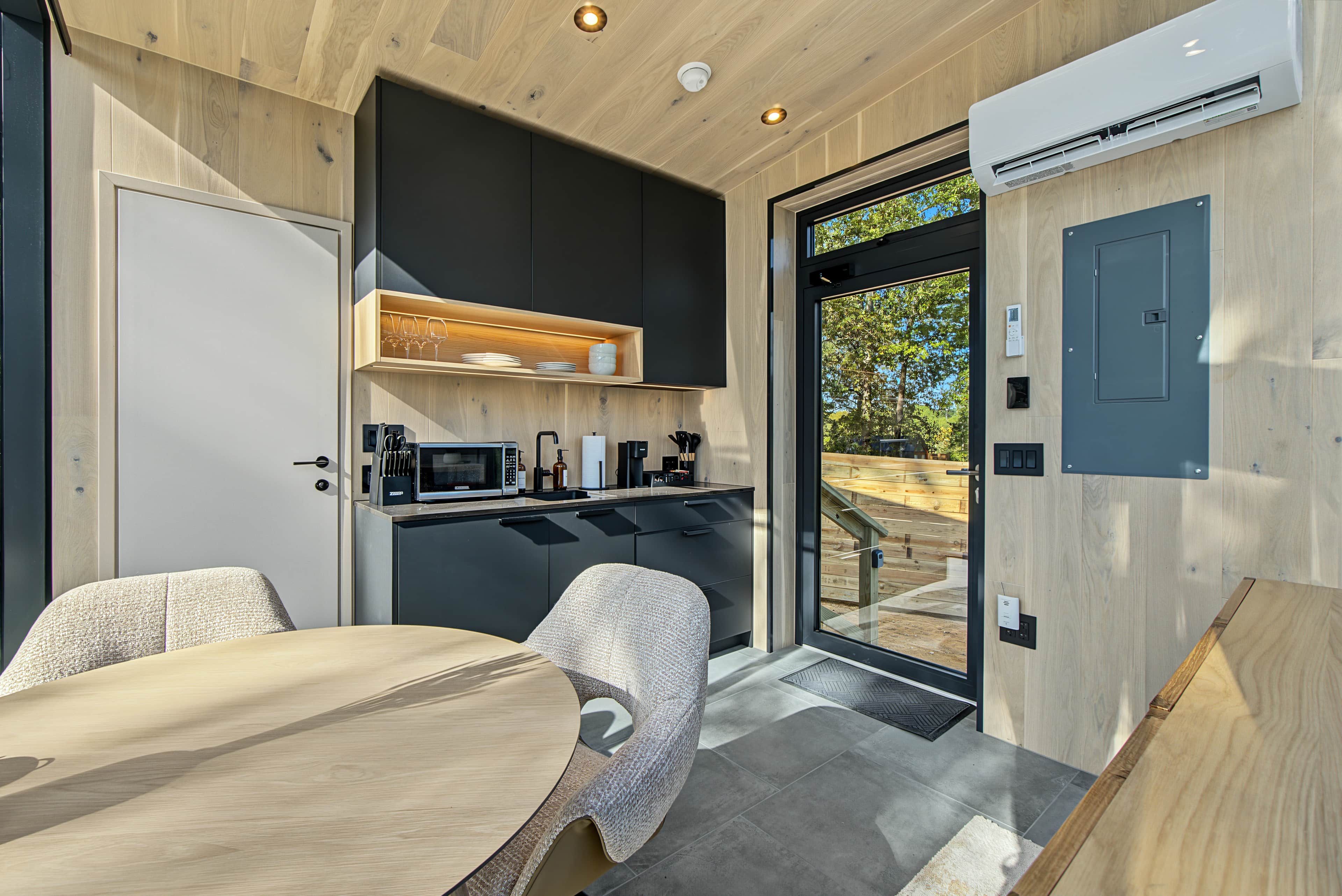 Een moderne keuken met een ingebouwde magnetron, koffiezetapparaat en een klein spoelbakje. Er staat een houten eettafel met stoelen, en een schuifpui naar een buitenruimte.