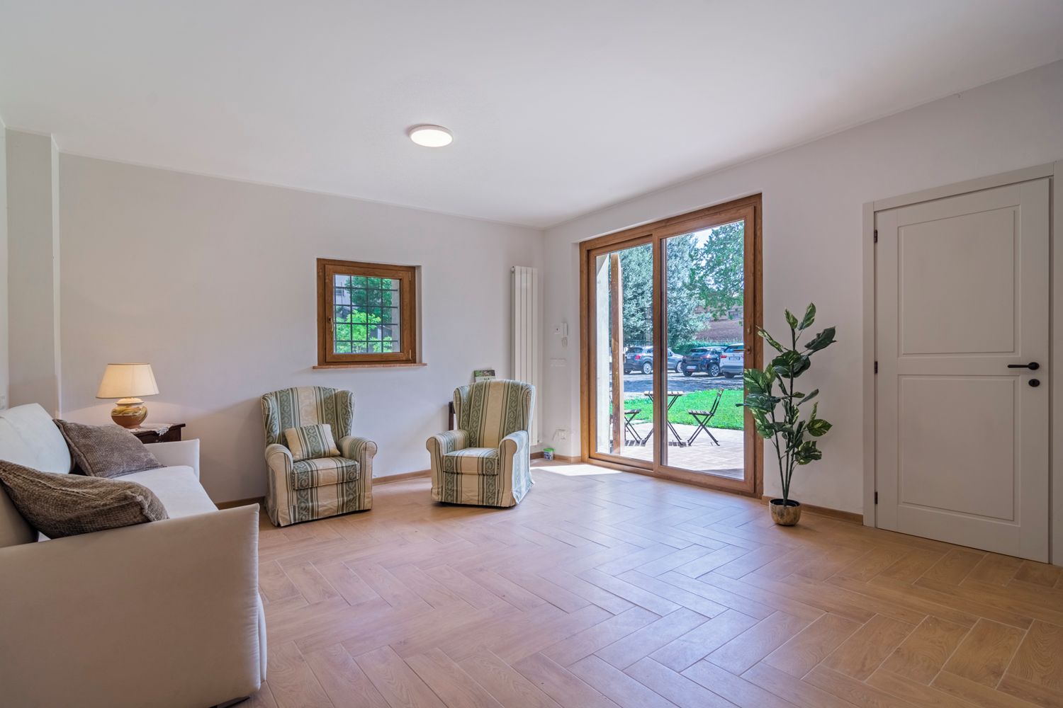 A spacious living room with a sofa, two armchairs, a lamp, a window, and a sliding glass door leading to an outdoor area.