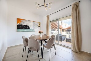 A dining area with a round table, six chairs, a vase with flowers, and a large abstract painting on the wall. - Gordonos – Modern 3Bed Apartment with Large Garden (Limassol)