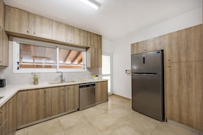 A modern kitchen with wooden cabinets, a stainless steel refrigerator, and a window with blinds. - Gordonos – Modern 3Bed Apartment with Large Garden (Limassol)